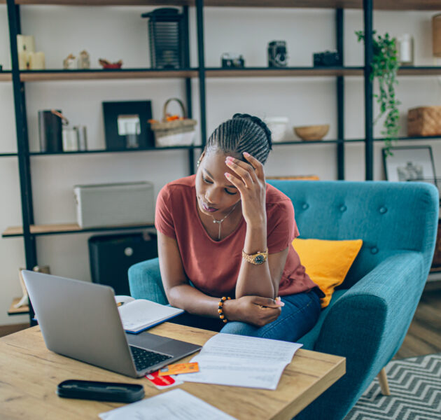 Young woman sitting at a table with laptop and notes. Her hand is placed on her head as if she is feeling stressed