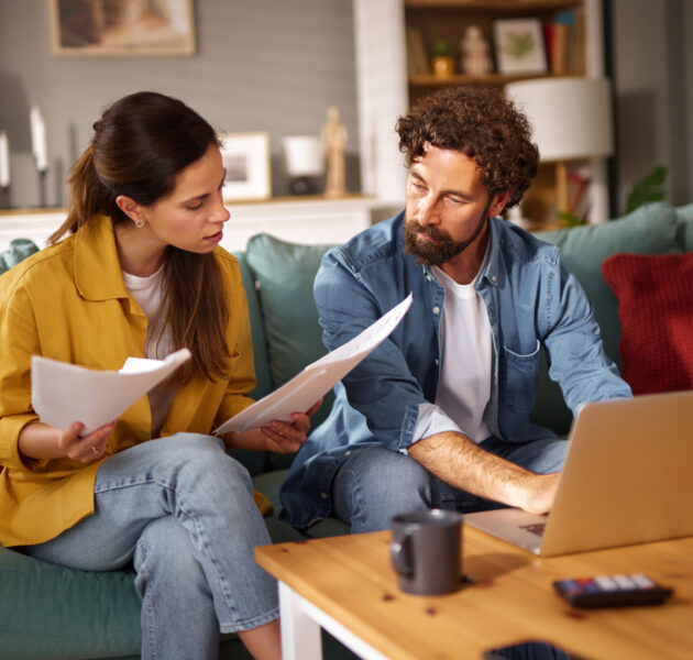 Two individuals are seated on a couch, focused on financial documents and a laptop, engaged in a serious conversation about their financial issues.