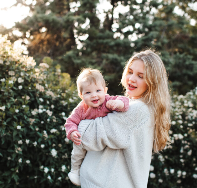 Smiling stylish woman holding baby girl 1 year old wearing knitted clothes over blooming bushes with flowers in meadow. Springtime. Motherhood.