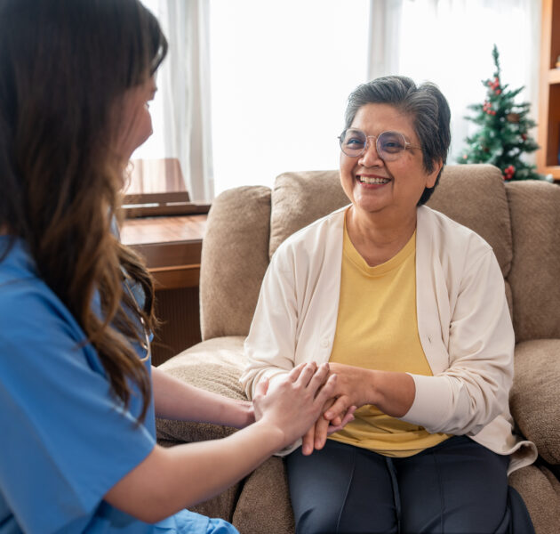 Smiling senior woman sitting on armchair holding hands with female caregiver for emotional support at home. Happy Asian elderly patient receiving professional nursing care and empathy in living room